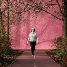 Older adult woman walking with mindful posture on a tree-lined path, eyes forward and arms relaxed, demonstrating focused gait and brain-body connection.